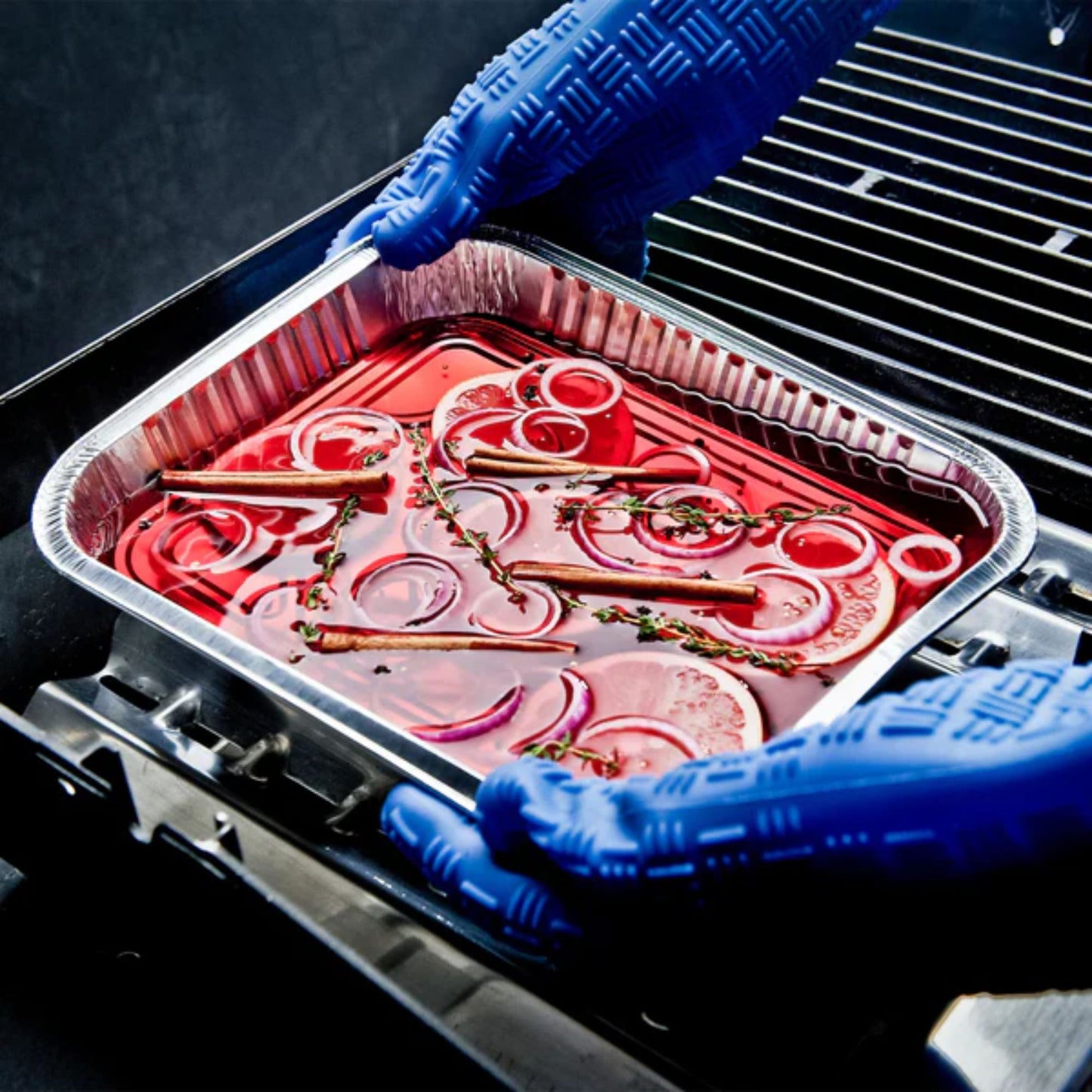 Person wearing blue gloves handling a tray of marinated meat and onions on a grill.