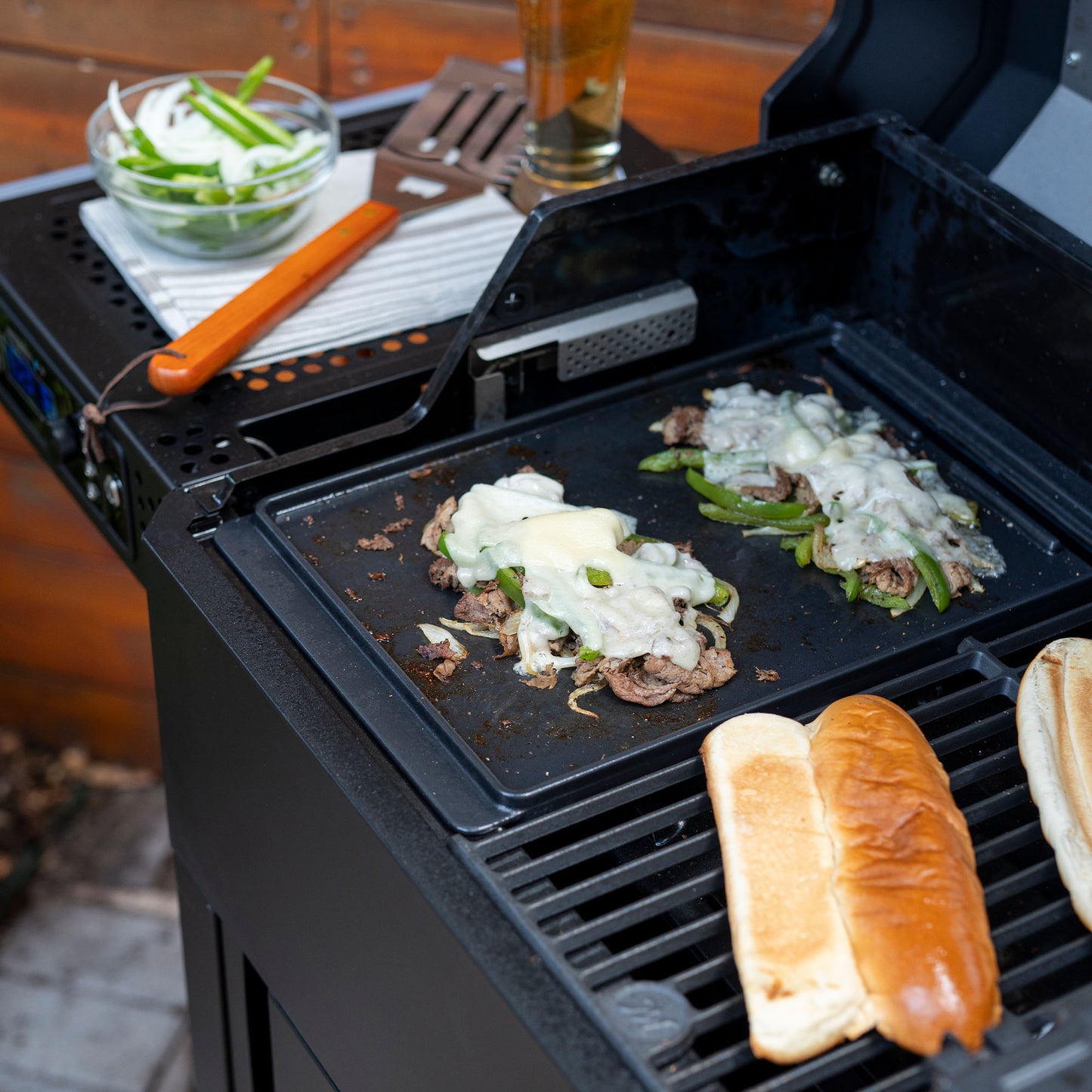 Grill with food being cooked, including a burger patty with cheese and vegetables, and buns.