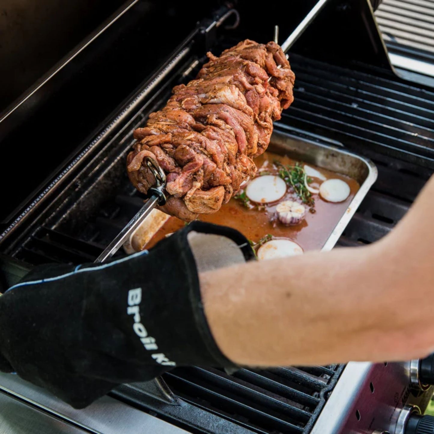 Person grilling a large piece of meat on a barbecue with a pan of vegetables.
