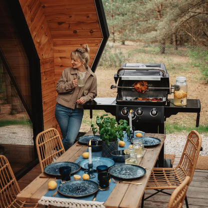A Broil King BBQ cooking with a women stood next to it and a table in front set ready for a meal. 