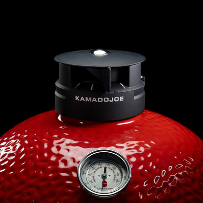 Red Kamado Joe grill with a black lid and thermometer on a black background