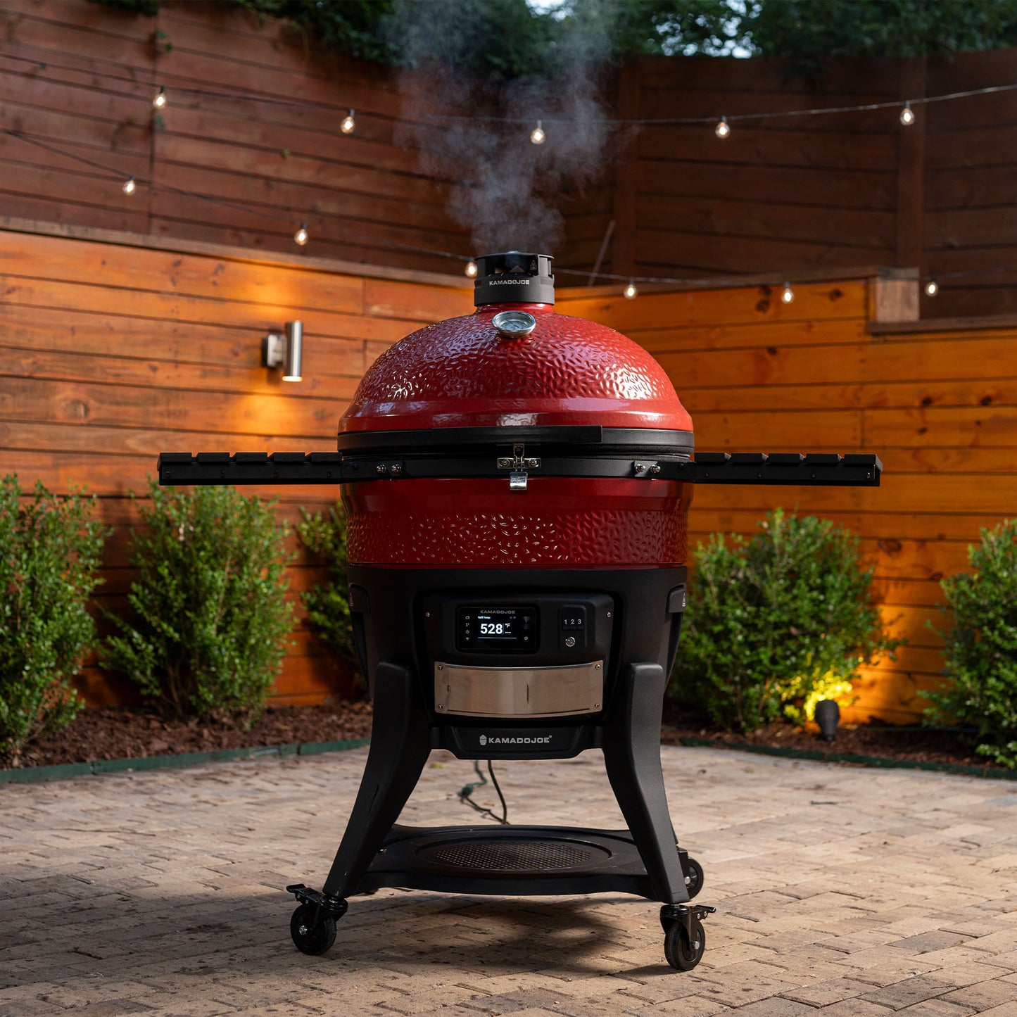 Red barbecue grill with smoke emitting from it, set against a wooden fence and outdoor setting.