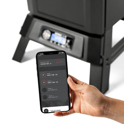 Hand holding a smartphone with Masterbuilt app interface in front of a 710 smoker.