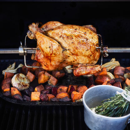 Roasted chicken on a rotisserie over vegetables with a cup of herbs on a grill.
