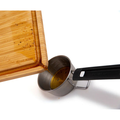 Wooden cutting board with a metal measuring cup containing oil on a white background