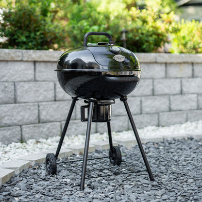 Black charcoal grill on a gravel surface with a stone wall and greenery in the background