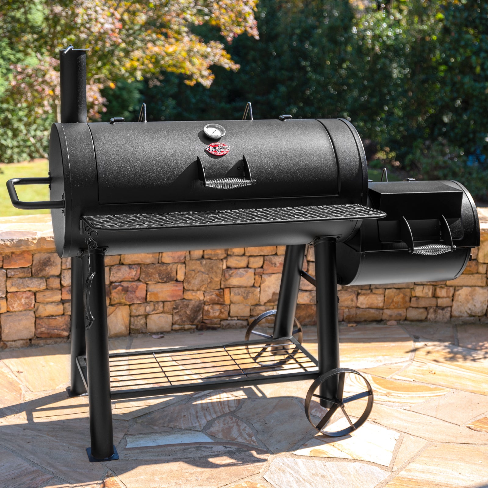 Black barrel-style barbecue grill on a patio with stone wall and greenery in the background