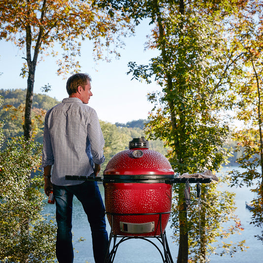 Man standing next to a red barbecue grill by a lake with trees in the background
