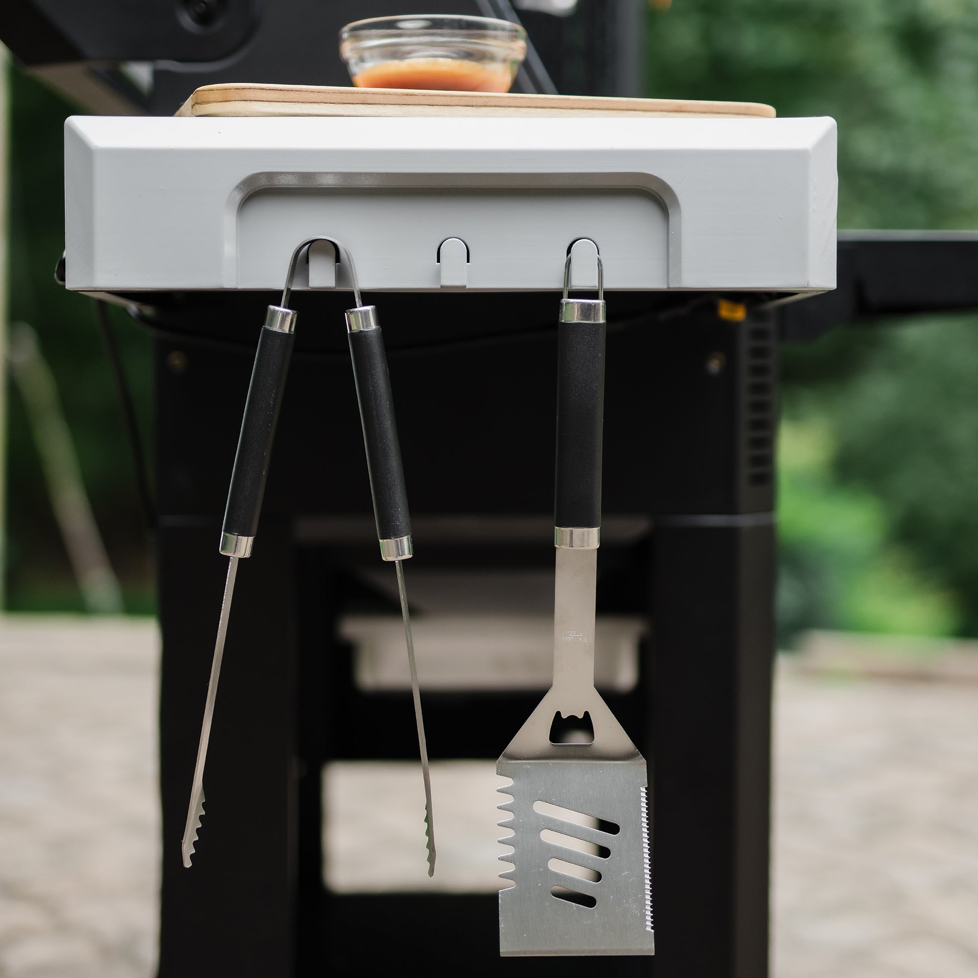 Barbecue tools hanging from a mounted holder on a grill with a blurred green background