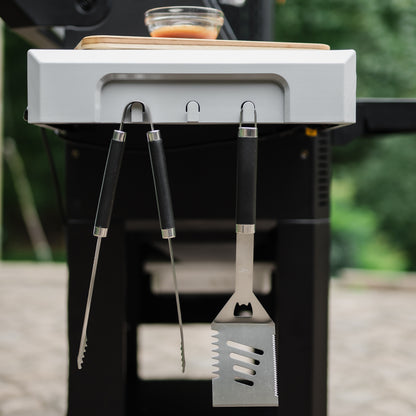 Barbecue tools hanging from a mounted holder on a grill with a blurred green background