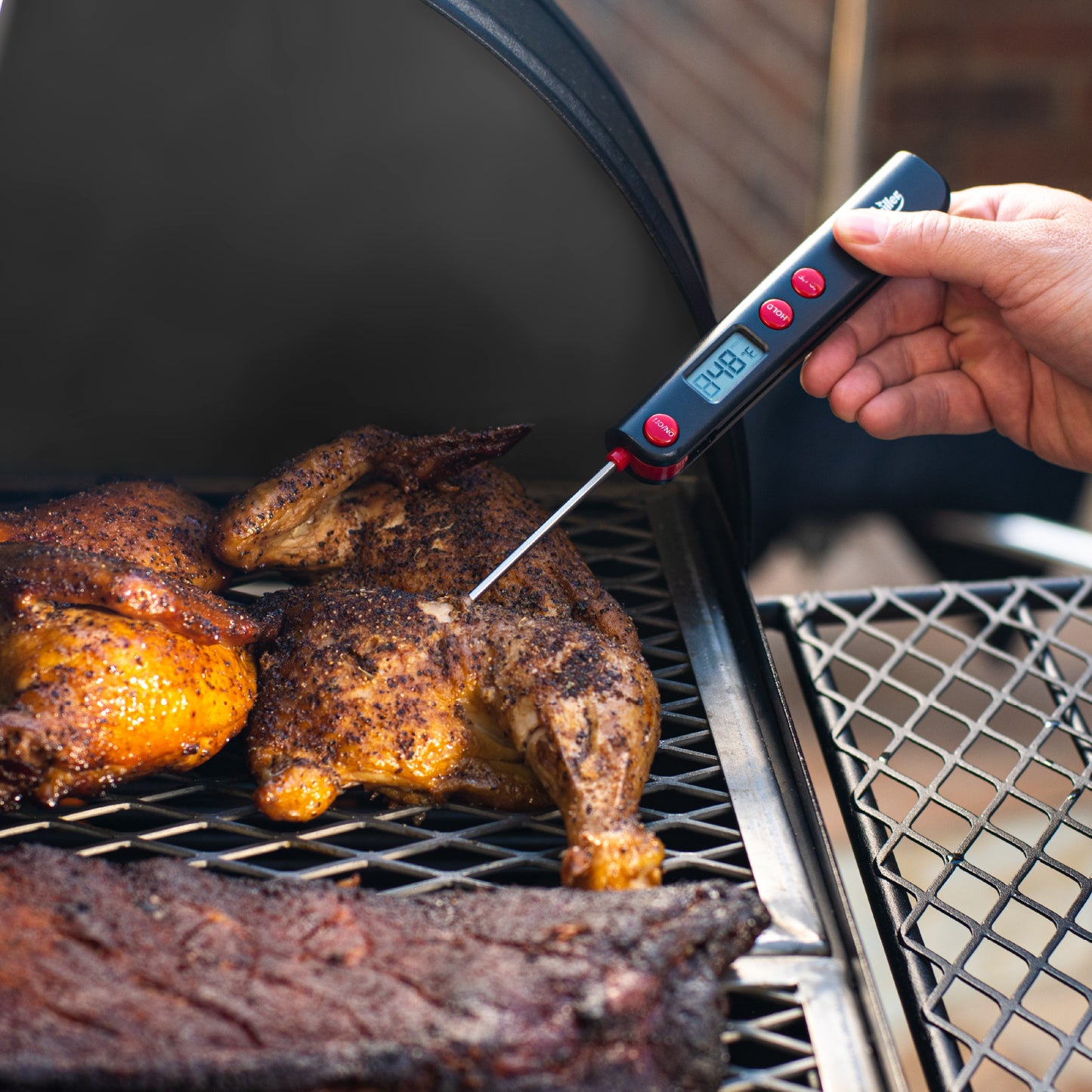 Person using a digital thermometer to check the temperature of roasted chickens on a grill.