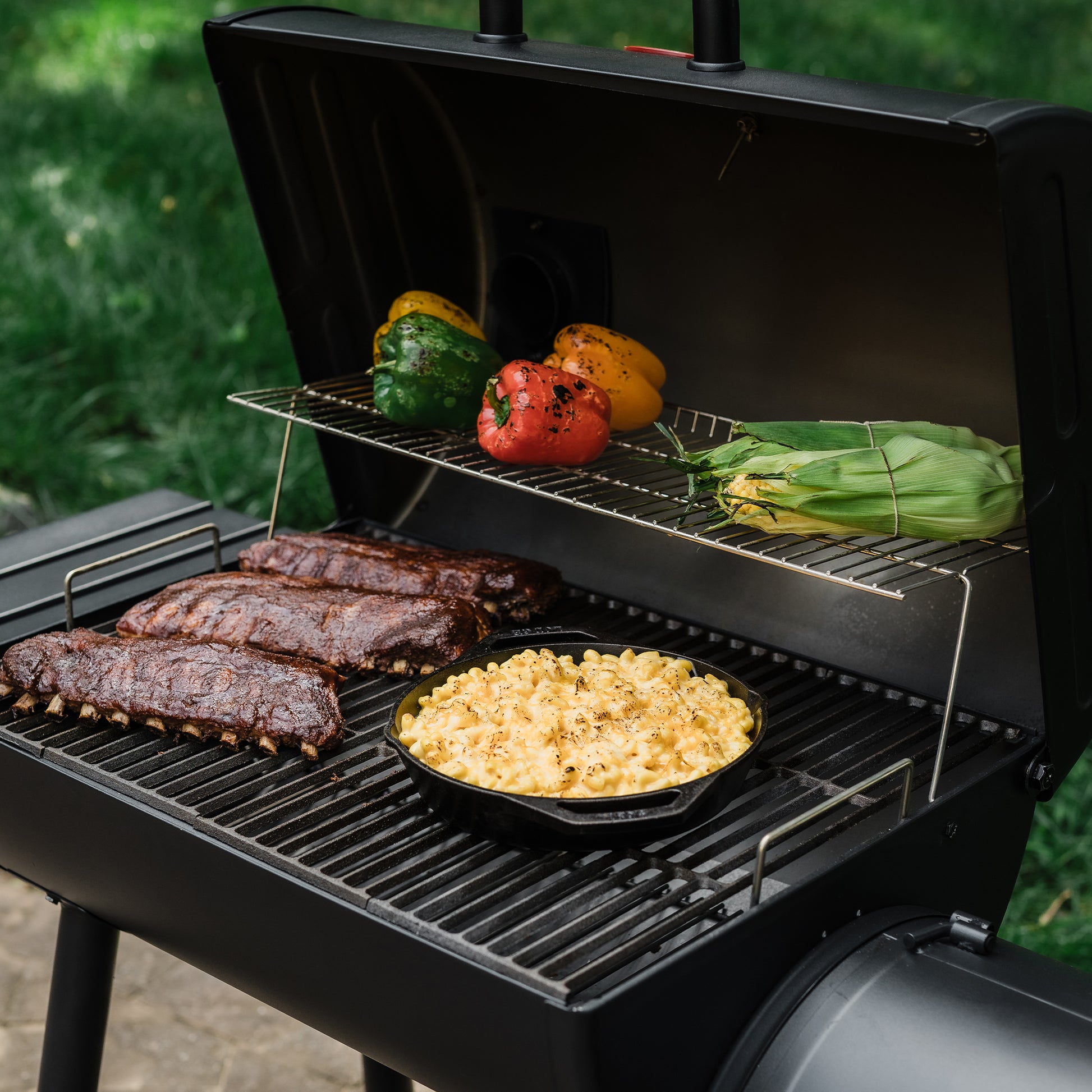 Grill with ribs, macaroni and cheese, and vegetables cooking on a metal rack.